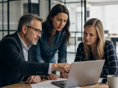Three business professionals collaborate around a laptop in a modern office, discussing cybersecurity risks.