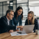 Three business professionals collaborate around a laptop in a modern office, discussing cybersecurity risks.