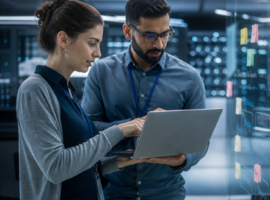 Two IT professionals reviewing data on a laptop in a modern server room