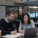 Two colleagues review supplier corrective action documents on a laptop in a modern office.